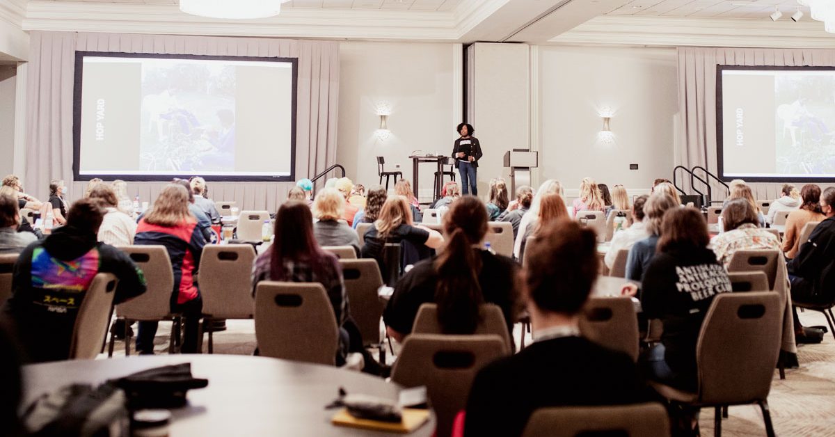 A room of people watch a woman speaking from a stage.